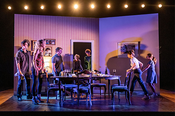 Seven people standing and spread out in a dining room under dim lighting