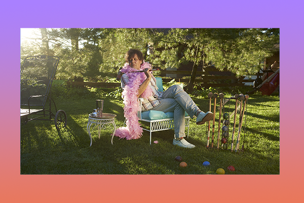 Ron Sexsmith wearing a feather boa sitting on a cushioned chair surrounded by trees