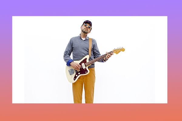 Hawksley Workman wears sunglasses, a sweater and corduroy pants, holding a guitar in a white void.